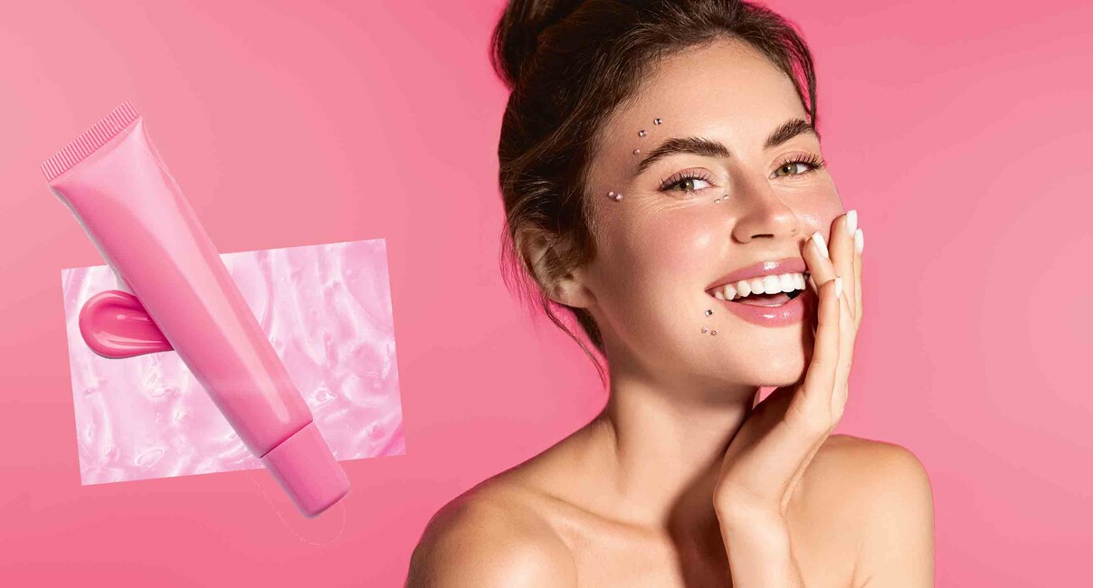Smiling woman with radiant, healthy skin on a soft pink studio background. Female model laughing, touching her flawless face without blemishes, has blush on her cheeks, standing with bare shoulders.
